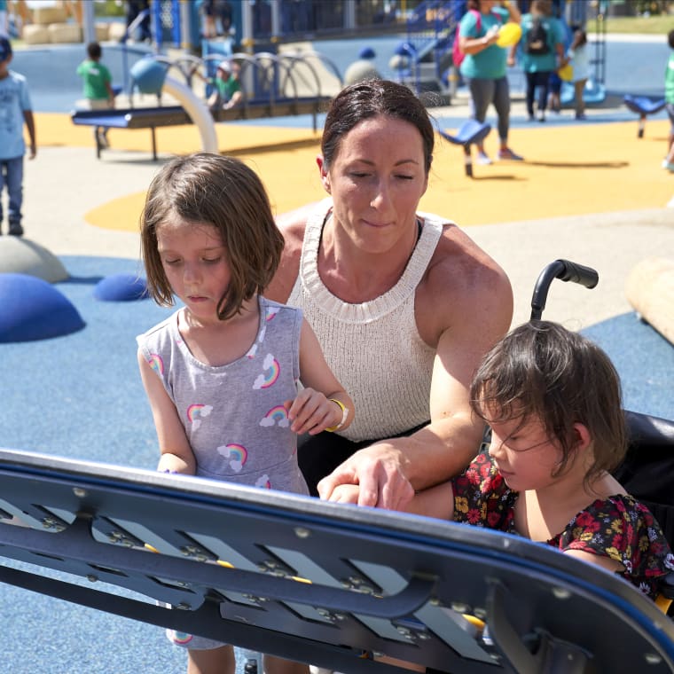 A woman helps two young girls, one seated in a wheelchair, interact with a low-mounted accessible play panel at a sunny, inclusive playground.