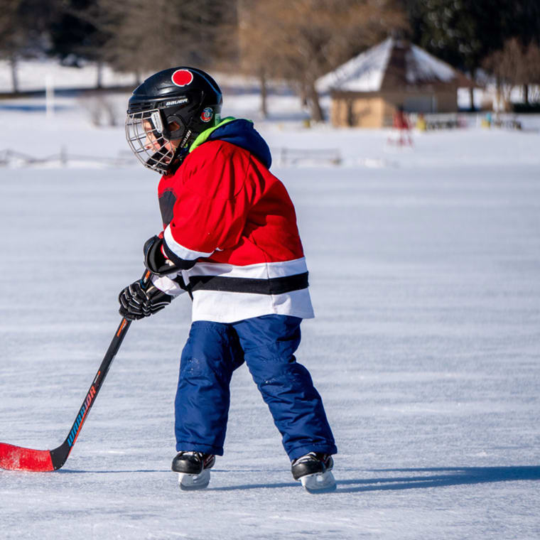 A child in a red hockey jersey, blue pants, and a black helmet stands on an outdoor frozen pond or lake, poised with a stick over a black hockey puck on a sunny winter day.