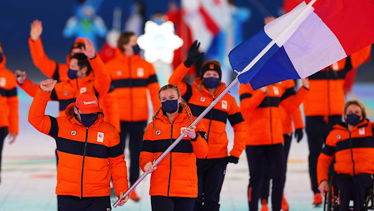 A group of athletes in orange Dutch kit marches out in the Opening Ceremony of the Paralympic Winter Games. The focus is on the two flagbearers at the top of the procession, a male and a female.