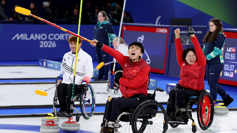Two wheelchair curlers celebrate after winning the game, the male pointing his broom and shouting while the female curler has both hands in the air.