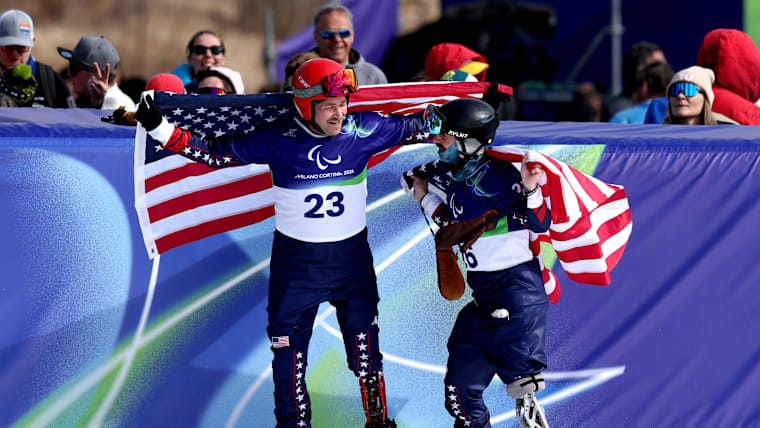 Two Para snowboarders with prosthetic legs showing hold up USA flags over their shoulders.