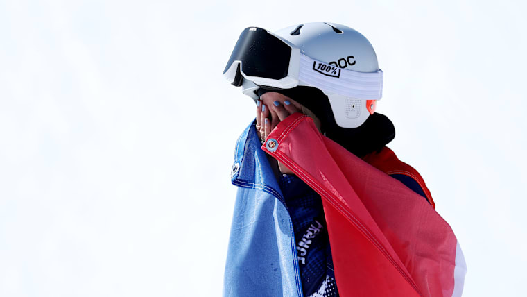 A female Para snowboarder in a white helmet and with a French flag draped over her shoulders covers her face with her hands in joy.