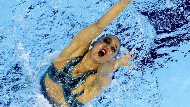 An overhead shot of a female artistic swimmer as she emerges from the water, her hands raised in the air.