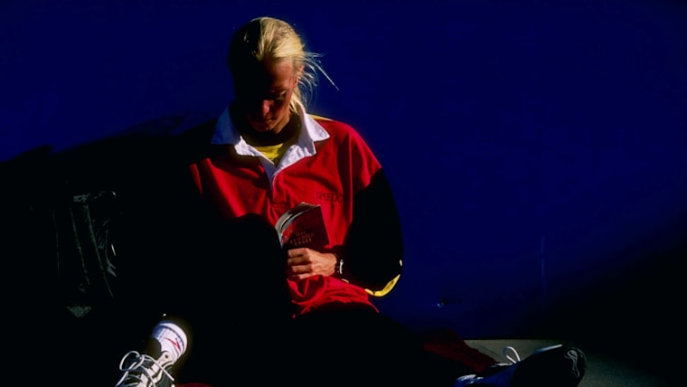 A female athlete reads a book while sitting on the floor of a competition venue.