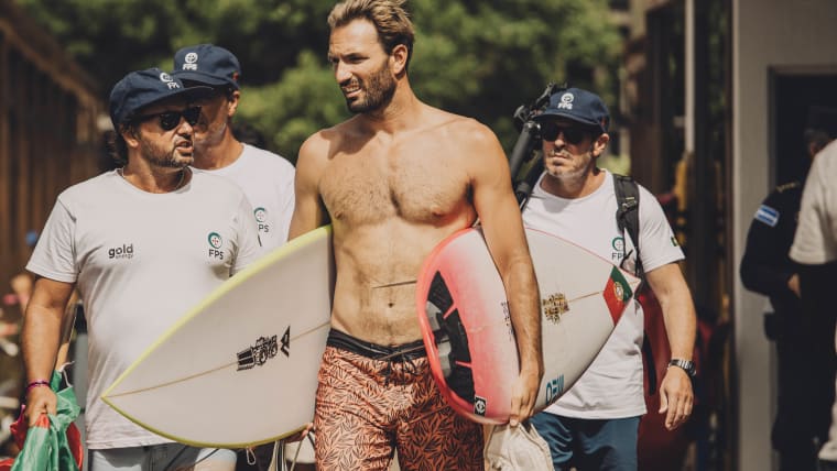 A male surfer holding two boards walks alongside his coaches.