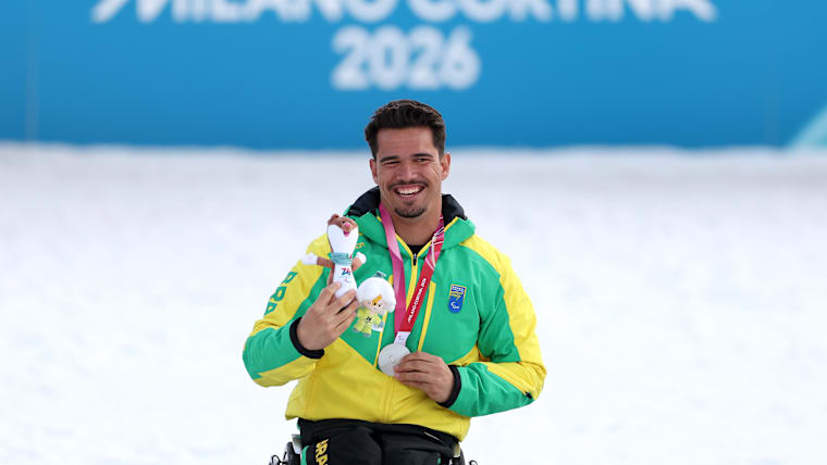 A Brazilian Para cross-country skier poses with his silver medal and mascot in front of a sign that says 'Milano Cortina 2026'.