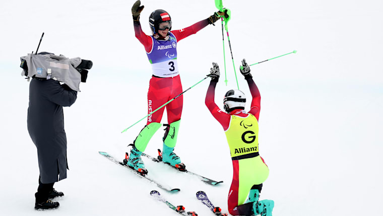 A male guide on his knees mock bows to an female Para Alpine skier, a cameraman on the left of the photo.