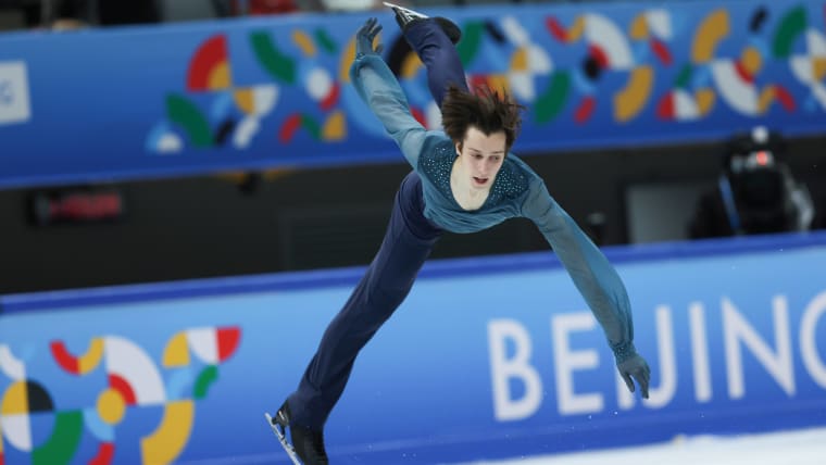 A male skater, dressed in a teal and black costume, enters a flying spin at a competition.
