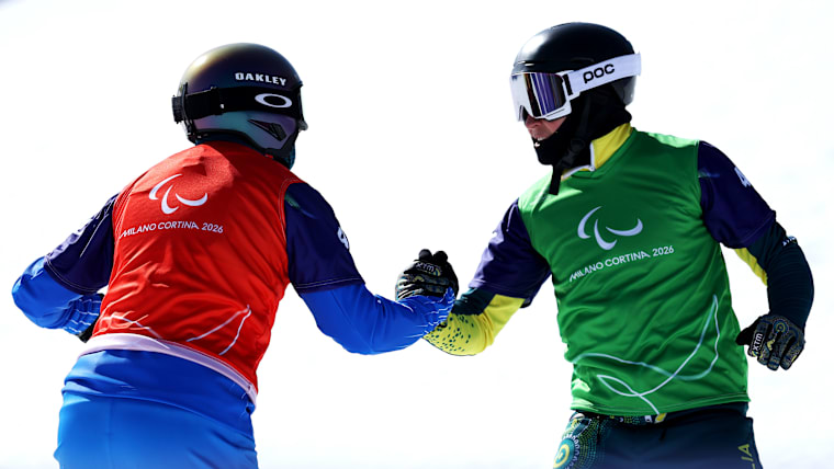 Two male snowboarders shake hands after crossing the finish line.