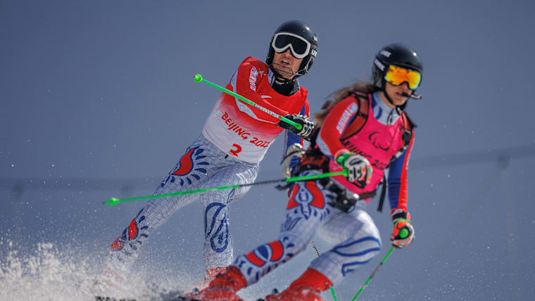 A male Para Alpine skier in black goggles follows a female guide down a mountain.