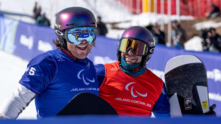 Two male Para snowboarders in helmets, bibs and goggles smile in the finish line.