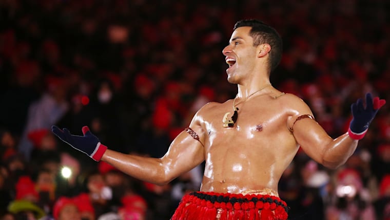 A shirtless male in traditional Pacific dress smiles and spreads out his arms on stage.
