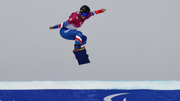 A female Para snowboarder soars over a snowboard cross roller during competition, the Agitos symbol imprinted on the snow.