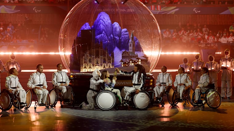 A girl in a wheelchair and a group of dancers in wheelchair around holds a golden torch, a large snow globe with Milan and mountain scenery blended in it, behind her.