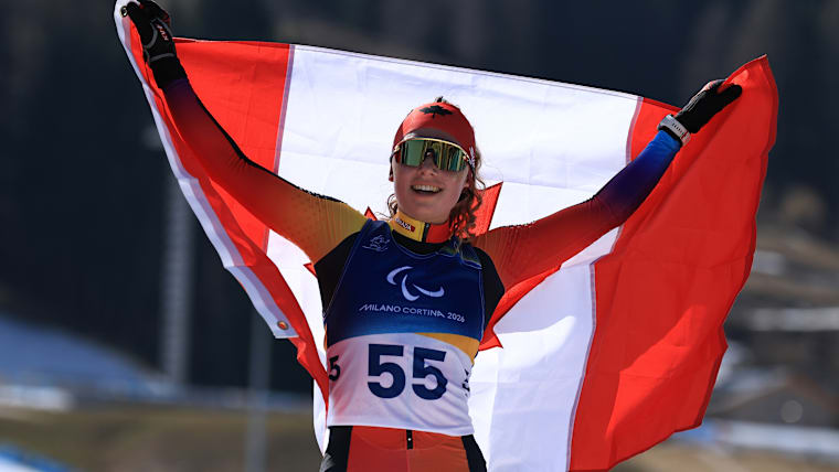 A Para biathlon athlete smiles while holding up the Canadian flag in celebration. 