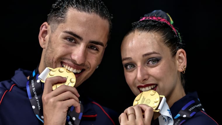 A male and female artistic swimmers bite their gold medals while posing on the podium.