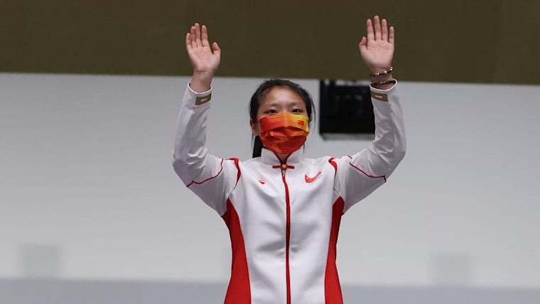 A female sports shooter raises her arms in celebration on the podium.