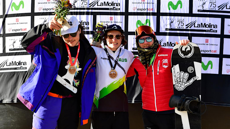 Three Para snowboarders pose in a medal ceremony, the one on the right making an alicorn sign with his hand and wearing a t-shirt with a unicorn on it.