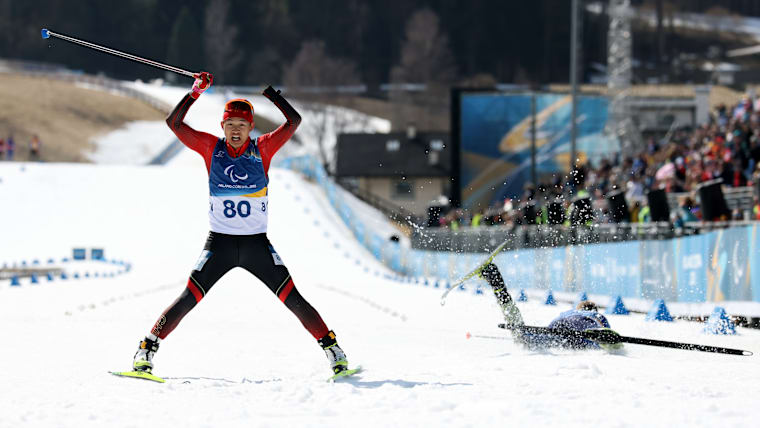 A Chinese Para biathlete celebrates with his arms stretched up as he crosses the finish line with his competitor falling to the ground in exhaustion.