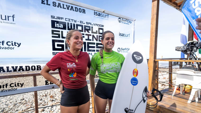Two female surfers pose by a clear board with World Surfing Games signage.