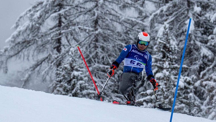 A male sit skier navigates a slalom course with trees behind him, the branches heaving from fresh snow.