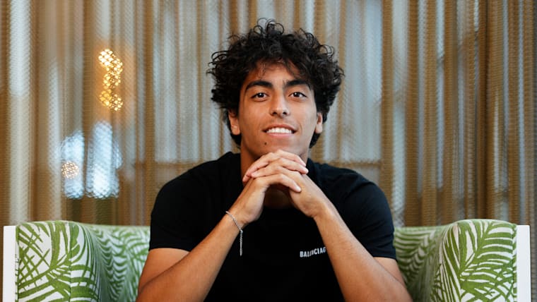 A seated Shelbayh smiles while posing for a photo with his fingers interlocked beneath his chin