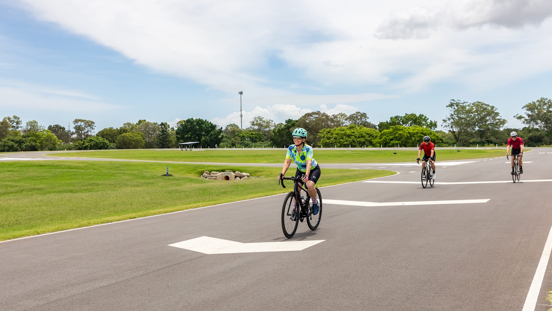 The sky is blue and the grass is green as three cyclist ride the circuit at Murarrie International Cycle Park