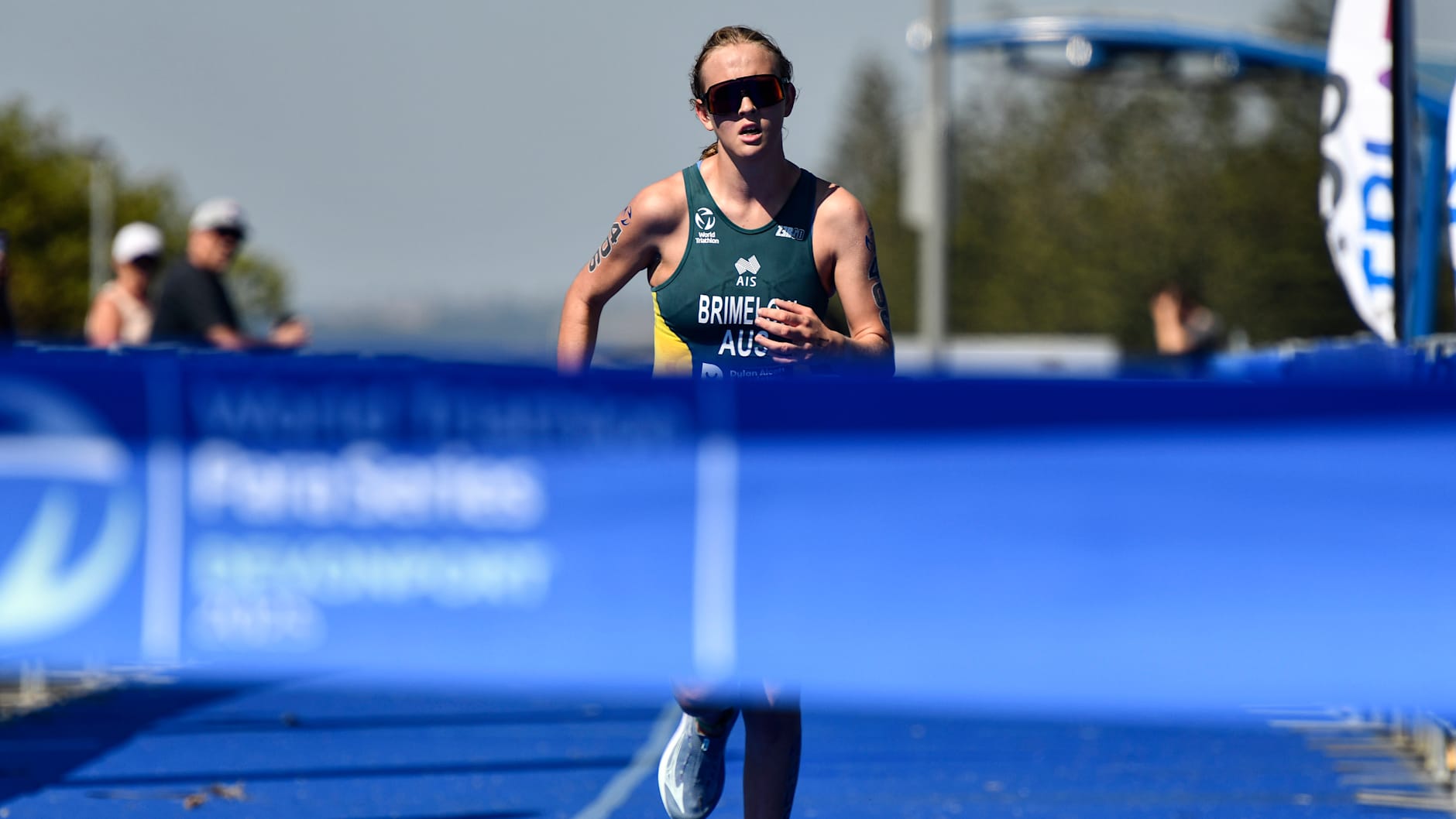 A young woman is about to reach the finish line of a race. She is wearing dark racing glasses and an Australian Paralympics uniform. 