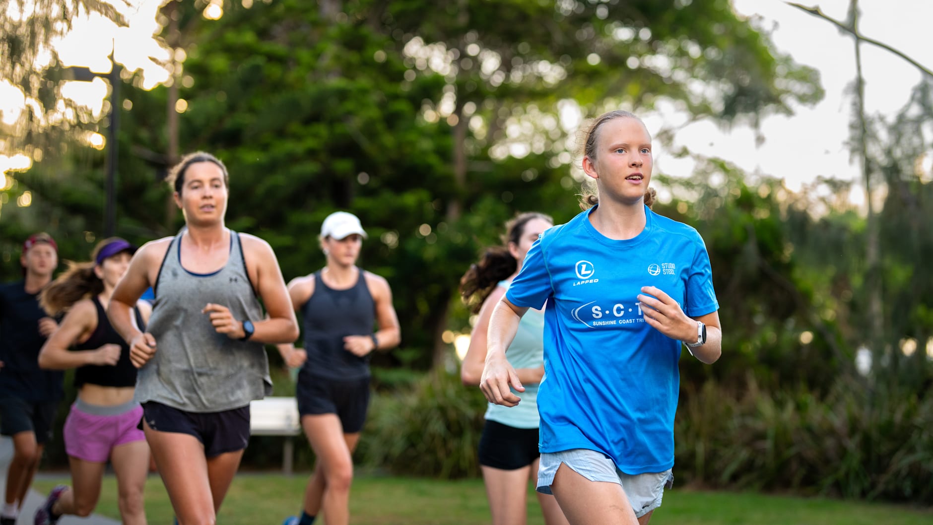 A group of runners are racing outdoors. Grace Brimelow is at the head of the group. She has blonde hair and is wearing a blue shirt. 