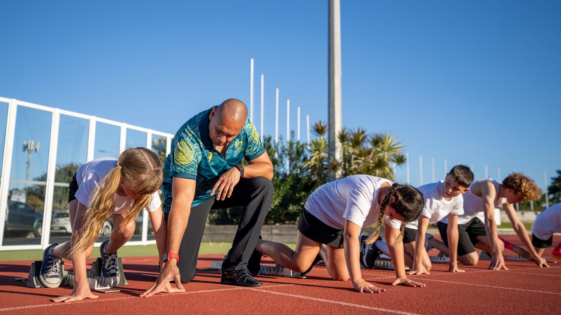 Patrick Johnson is at the starts line of the 100m with several kids in a white shirt demonstrating how to do the perfect start