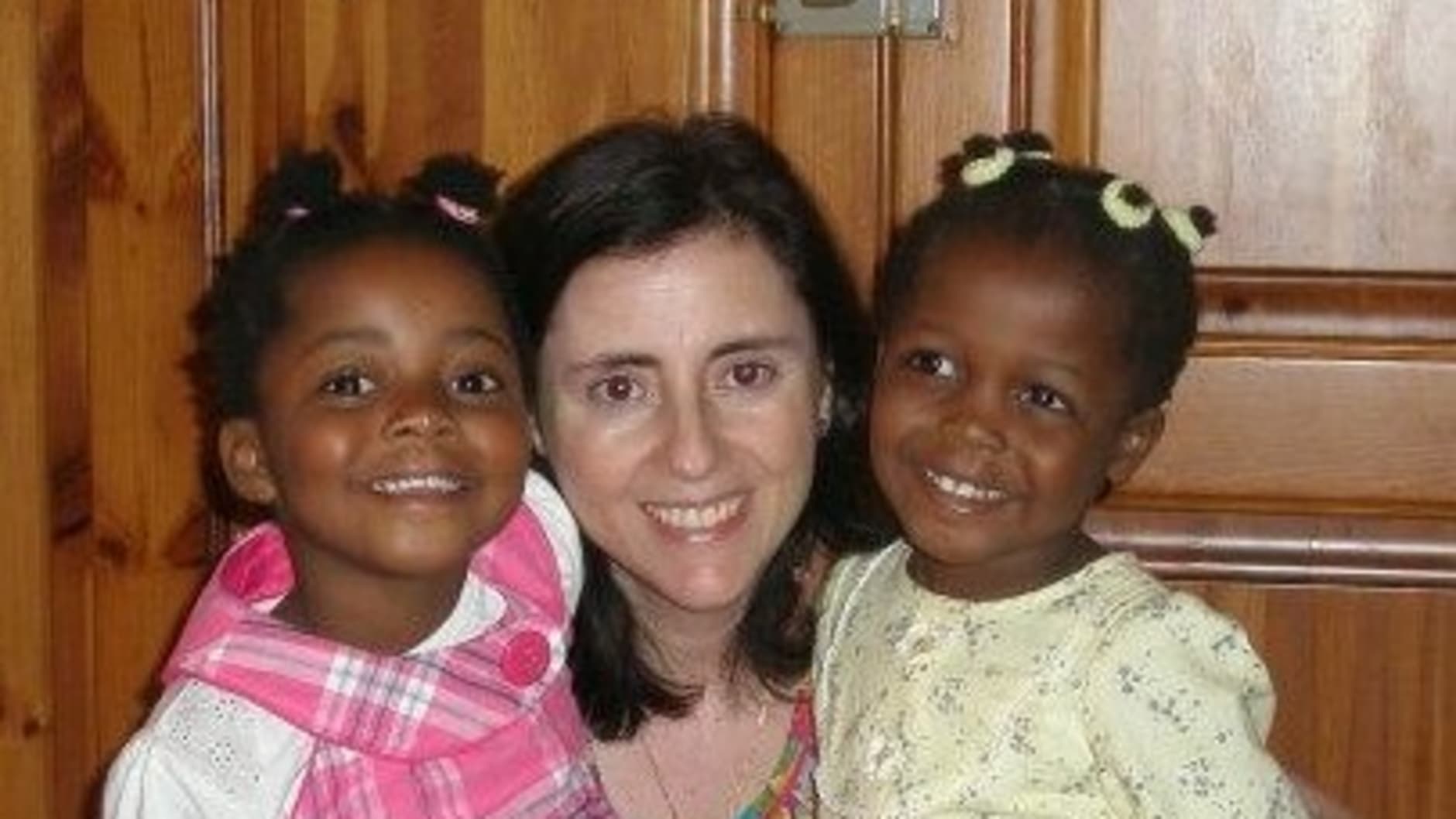 Two young girls of colour, one dressed in pink tights and a dink checkered dress and the other in white tights and a pale yellow dress with long sleeves, with their hair tied up pose for a photo with their mum, who is wearing jeans and a colourful top.