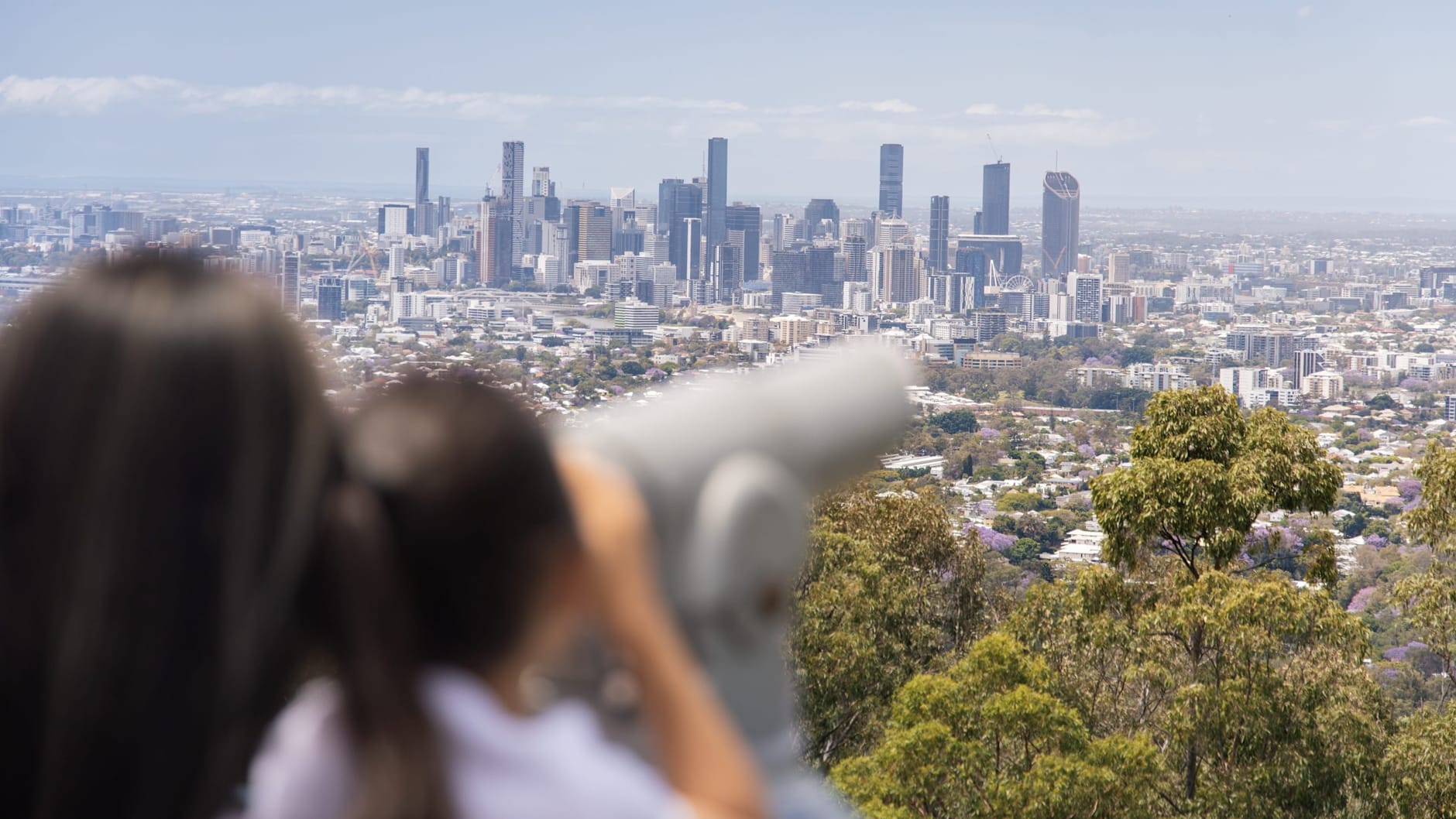 Two people blurred in the foreground are looking through binoculars out over Brisbane City from the Mt Coot-tha vantage point.