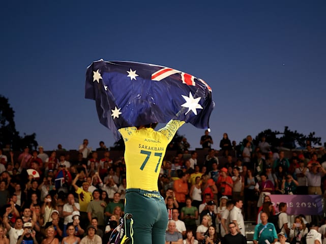 A photo from behind shows Saya Sakakibara raising the Australian flag above her head with her BMX bike below her as she stands on a hill.