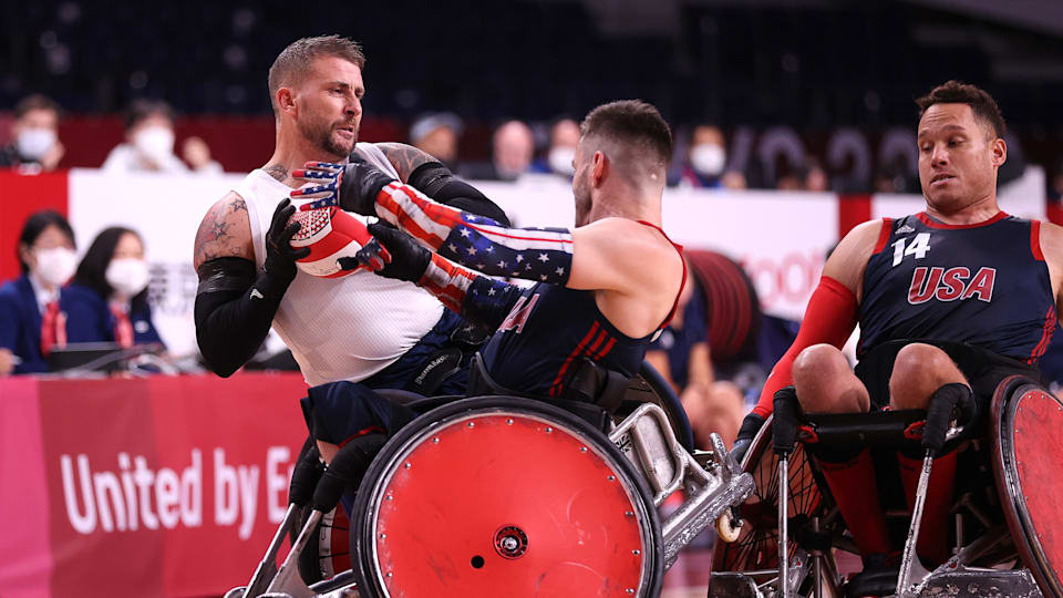 Two male wheelchair rugby players compete for the ball while another looks on.