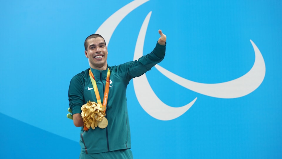 A male swimmer with a gold medal around his neck and an Agitos sign in the background waves to the spectators during a victory ceremony.